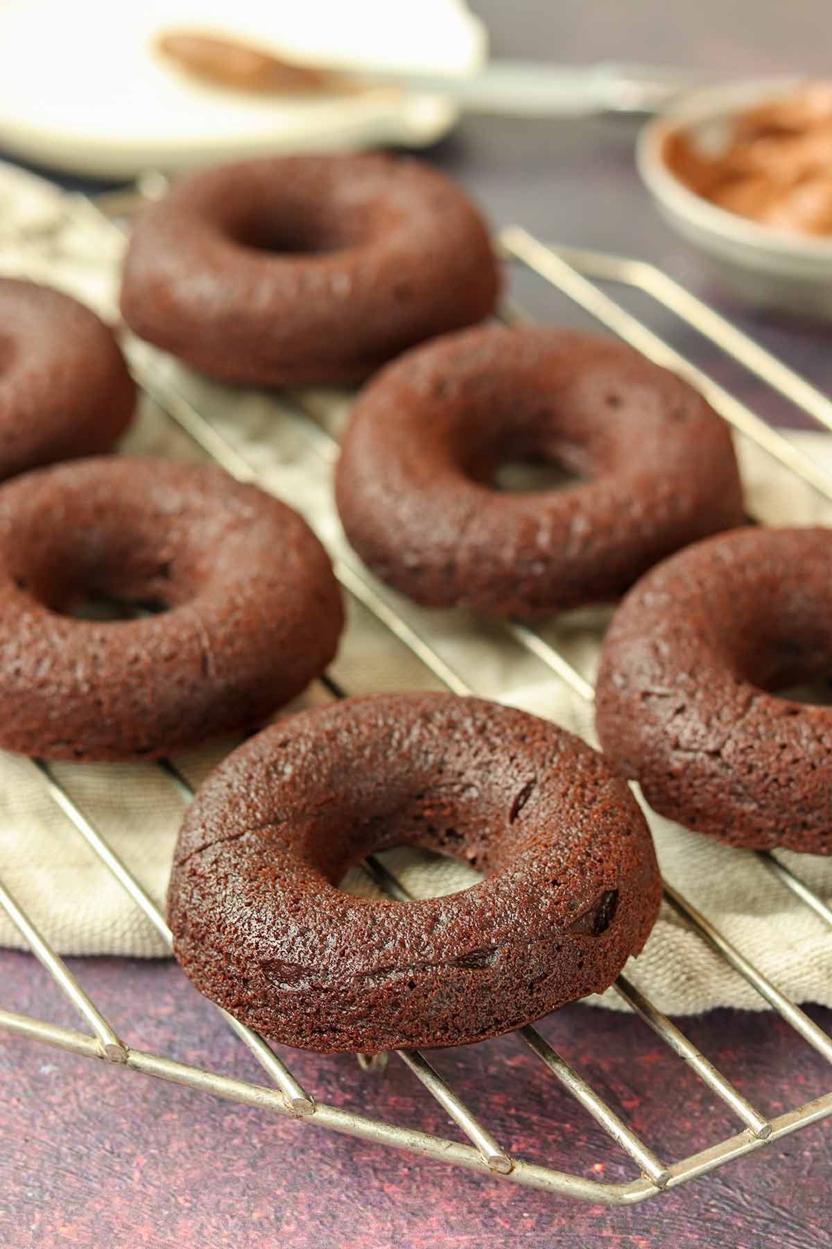 Chocolate cassava flour donuts lined up on a wire cooling rack surrounded by ingredients.