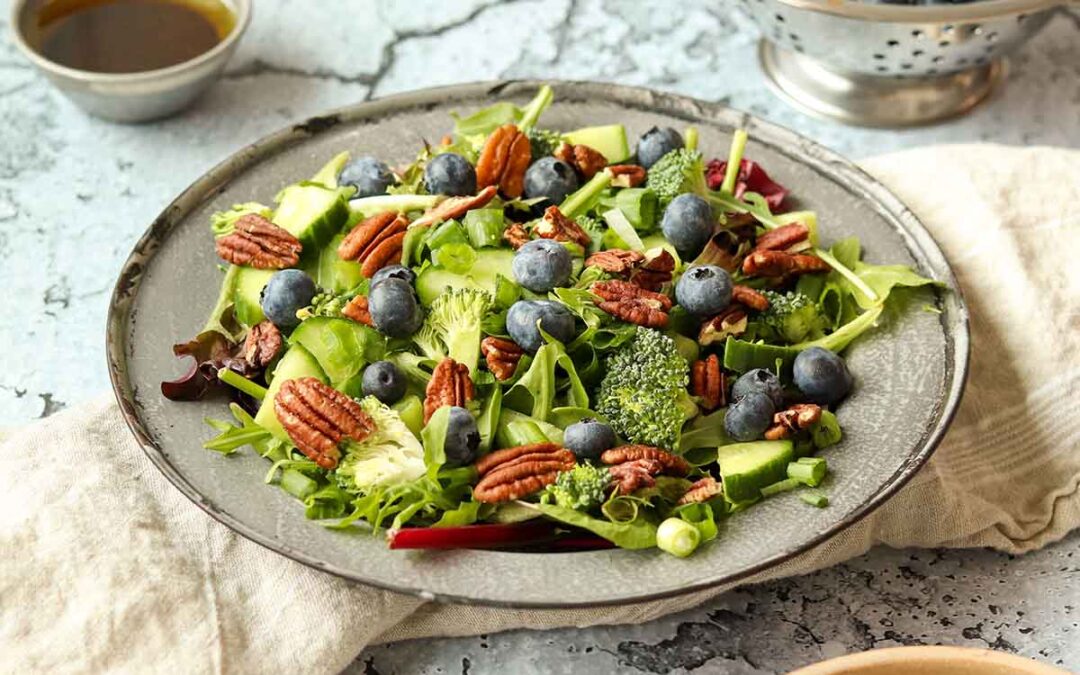 Side view of Summer pecan salad surrounded by a colander of blueberries and a metal dish filled with vinaigrette.