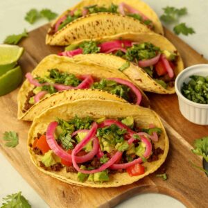 Close up side view of bison tacos lined up on wooden serving platter surrounded by brightly coloured garnish.