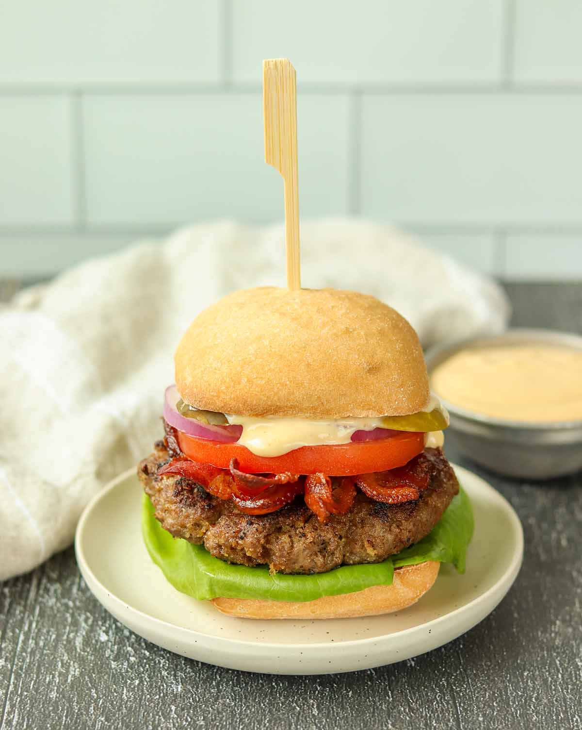 Close up overhead view of moose burger on white ceramic plate surrounded by a white dish cloth and a ramekin of sauce.