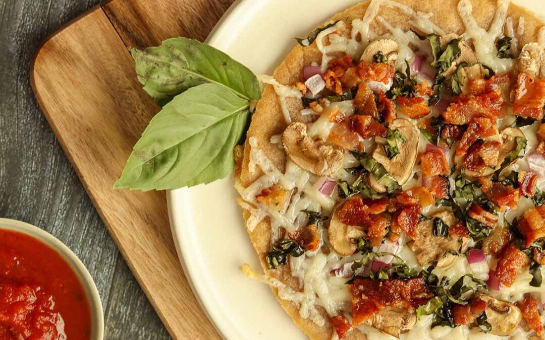 Close up side view of cassava flour pizza on a wooden serving board surrounded by sauce and fresh basil.