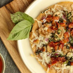 Close up side view of cassava flour pizza on a wooden serving board surrounded by sauce and fresh basil.