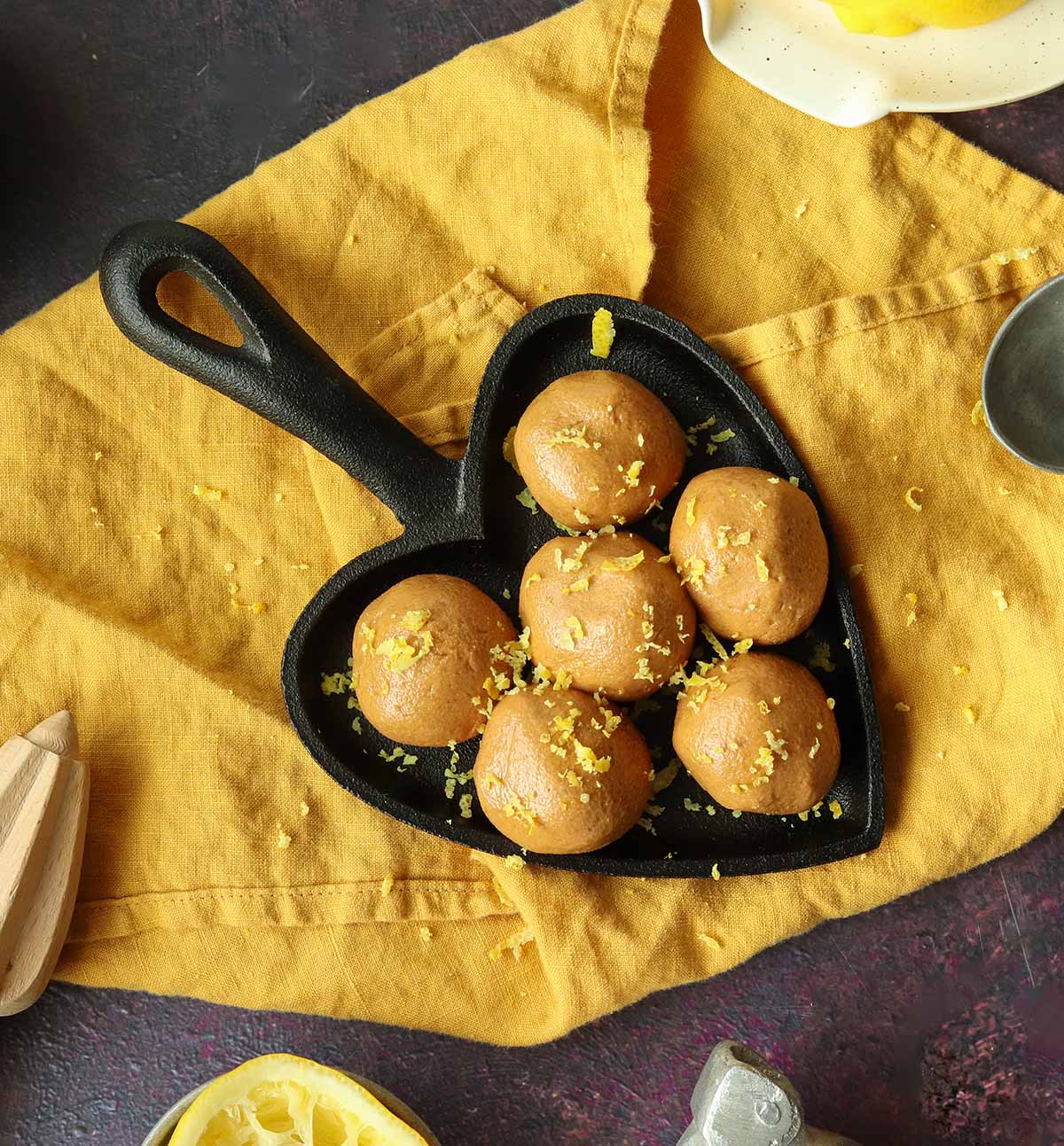 Overhead view of collagen balls in a heart shaped skillet placed onto a yellow kitchen towel surrounded by ingredients and sprinkled with lemon zest and shredded coconut.